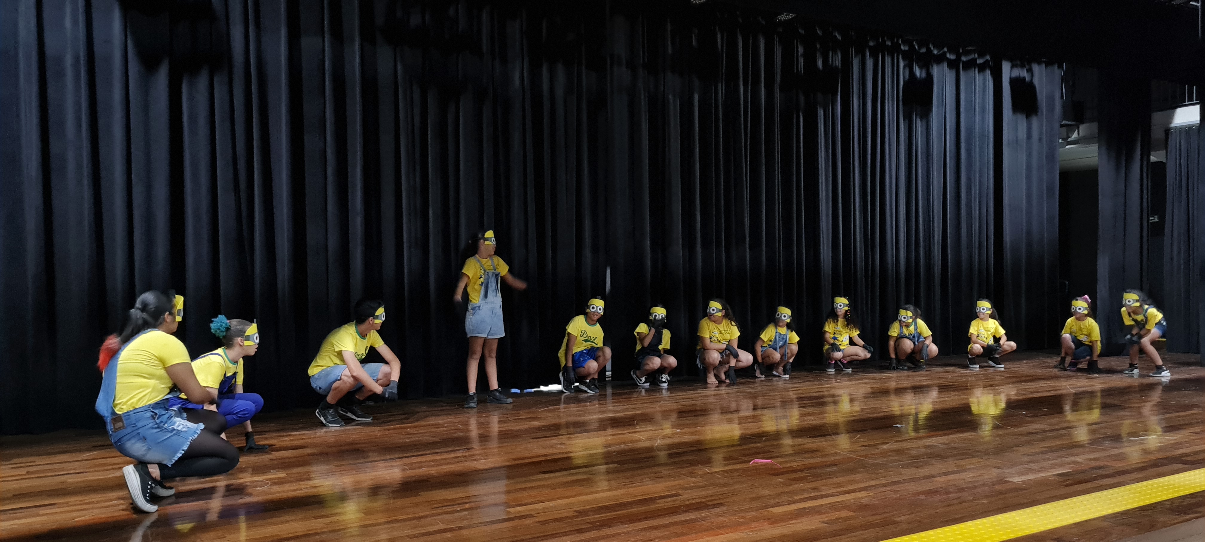 Com roupas amarelas e usando óculos, crianças estão no palco do auditório do CEU Freguesia do Ó realizando apresentação para plateia.